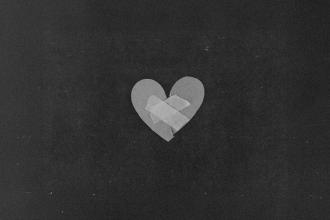 A simple paper heart sits against a dark background with two pieces of tape placed in an “X” shape across the center