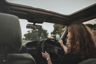 A person drives a car with the roof open, holding a phone in one hand while steering with the other
