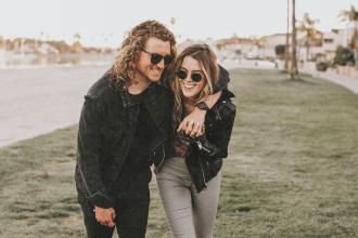 Two people walk arm‑in‑arm across a grassy area near a waterfront path, dressed casually in jackets as they lean affectionately toward each other