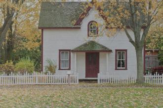 A small white house with red trim sits behind a white picket fence, surrounded by tall trees with golden autumn leaves scattered across the yard