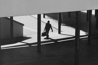 A silhouetted figure walks through a modern outdoor walkway carrying a bag, with strong shadows cast across the ground