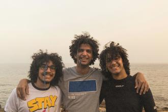 Three friends stand arm in arm near the shoreline, with the sea and sky forming a soft backdrop