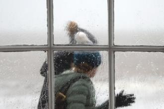 Two people in winter clothing walk past a rain‑covered window, their figures softened by the water droplets on the glass