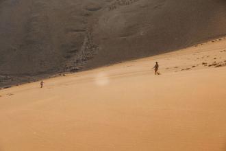 Two people walk across a vast desert slope, appearing small against the large sandy landscape