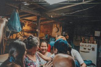 Several people gather around someone in a rustic, dimly lit shelter, placing comforting hands on their shoulders