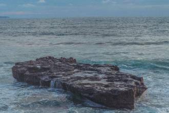 A large flat rock formation sits in the ocean as waves wash gently over its edges under a soft blue sky