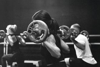 A group fitness class performs barbell squats, with participants lifting weighted bars across their shoulders