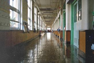 A long, empty school hallway lined with windows and classroom doors reflects bright light on its shiny floor