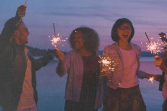 A group of people stand together at dusk near the water, holding sparklers that glow against the purple sky