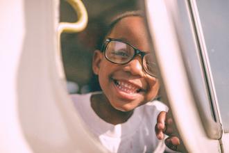 A child sits by a window on a vehicle, resting a hand on the window frame while looking outside