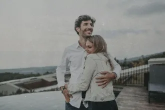 Two people stand together outdoors on a rooftop or terrace, with one person’s arm wrapped around the other as they look toward the landscape