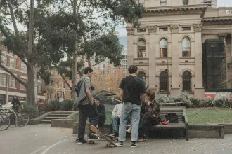 A group of people gather in an outdoor plaza near a bench, with skateboards nearby and historic buildings in the background