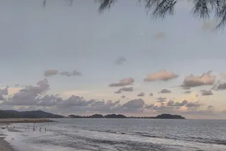 A calm beach scene at dusk with soft waves, scattered clouds, and distant tree‑covered hills across the shoreline