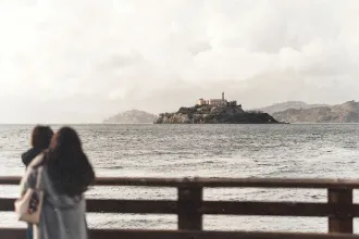 Mother holding her child, standing on a wooden pier looking across the water toward an island with historic stone buildings