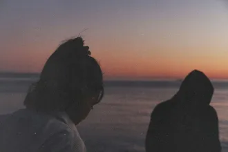 Two people stand near the water at sunset, their silhouettes softly outlined against a warm orange and purple sky