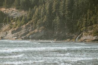 Gentle ocean waves roll toward a forested coastline with steep rocky cliffs and a small waterfall flowing into the sea