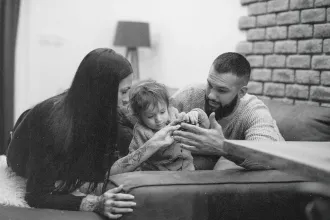 Two adults and a small child sit together on a couch, leaning in as they help the child with something in their hands