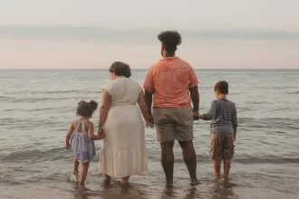 A family of four stands together at the edge of the ocean, holding hands as gentle waves wash over their feet during a calm sunset
