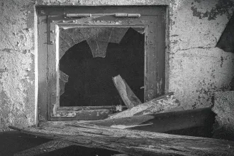 A broken window with shattered glass sits in a weathered concrete wall surrounded by debris