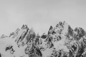 Sharp, jagged mountain peaks covered in snow rise dramatically into a pale, overcast sky, with patches of mist drifting around the ridges