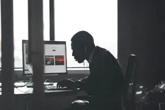 A person sits at a desk working on a computer with large windows behind them, silhouetted against the light