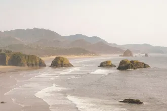 Ocean waves roll toward a sandy shoreline with large rocky sea stacks and distant mountains under a hazy sky