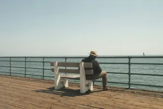 A person wearing a hat sits alone on a wooden pier bench, looking out over pier railing at a calm ocean