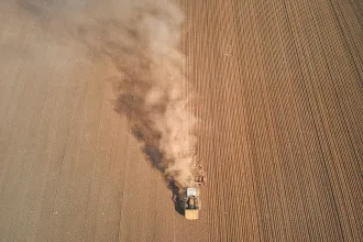A tractor moves across a large plowed field, leaving a cloud of dust trailing behind it