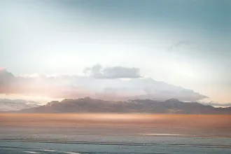 A distant mountain range sits beneath soft clouds as warm light spreads across a wide, flat desert foreground