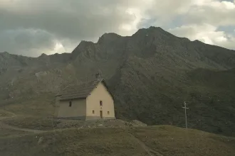 A small church sits on a grassy hillside beneath towering, rugged mountains under a cloudy sky