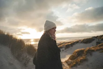 A woman wearing a knit beanie and coat walks along sandy dunes near the beach at sunset, with warm light shining through clouds over the ocean