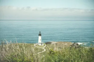 A white lighthouse stands on a grassy cliff overlooking calm blue ocean water, with clouds lining the distant horizon
