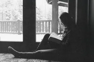 A person sits on the floor beside large glass doors, reading a book while snow-covered trees and a porch are visible outside