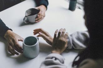 Three hands resting near ceramic mugs on a white tabletop, suggesting conversation over drinks