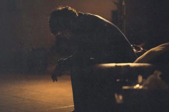 A person sits leaning forward on a couch in a dimly lit room, illuminated by warm, low light