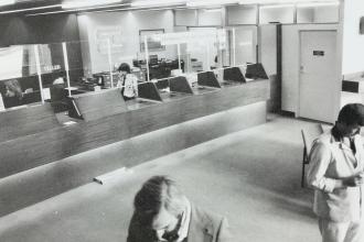 Interior of a bank lobby with teller counters, glass partitions, and several people moving through the space