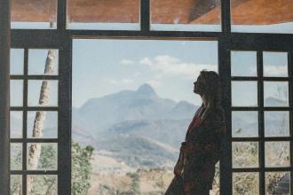 A person stands in profile inside a large window frame, looking out toward distant mountains under a bright sky