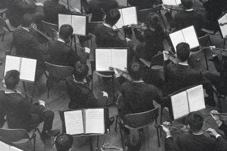 Musicians seated in rows playing string instruments with open sheet music in a formal rehearsal setting