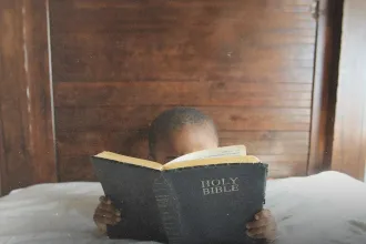 Young child holding and reading an open Bible while sitting indoors against wooden paneling