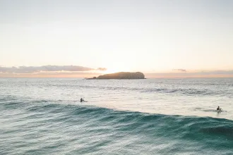 Two surfers sitting on their boards in gentle ocean waves at sunrise with a small island in the distance
