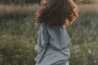 Person with curly hair standing in a grassy field with warm sunlight shining from behind
