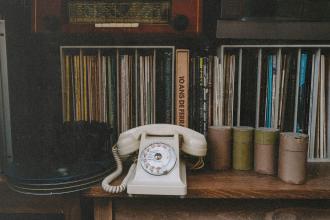 Vintage rotary telephone displayed on a wooden shelf surrounded by vinyl records, books, and small storage canisters