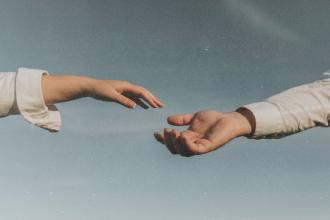 Two outstretched hands reaching toward each other against a soft blue sky, as if about to connect