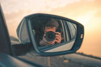 Photographer holding a camera reflected in a car’s side‑view mirror during a warm sunset drive