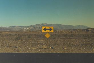 Yellow road sign with arrows pointing left and right in a barren desert landscape with distant mountains under a hazy sky