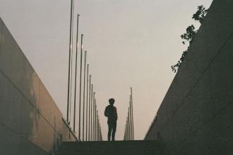 Silhouetted figure standing at the top of a walkway lined with tall flagpoles under a hazy evening sky
