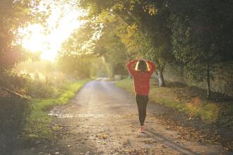 Person walking down a sunlit tree‑lined path at sunrise, wearing a red sweater and stretching with arms raised overhead