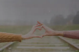 Two people extending their arms to form a heart shape with their hands over blurred railroad tracks