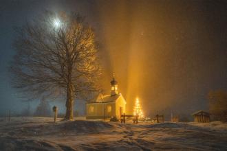 Snow‑covered church glowing with warm golden lights at night, with a lit Christmas tree and a large bare tree under a bright moon