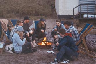 Group of friends sitting around a small beach bonfire, chatting and relaxing in deck chairs